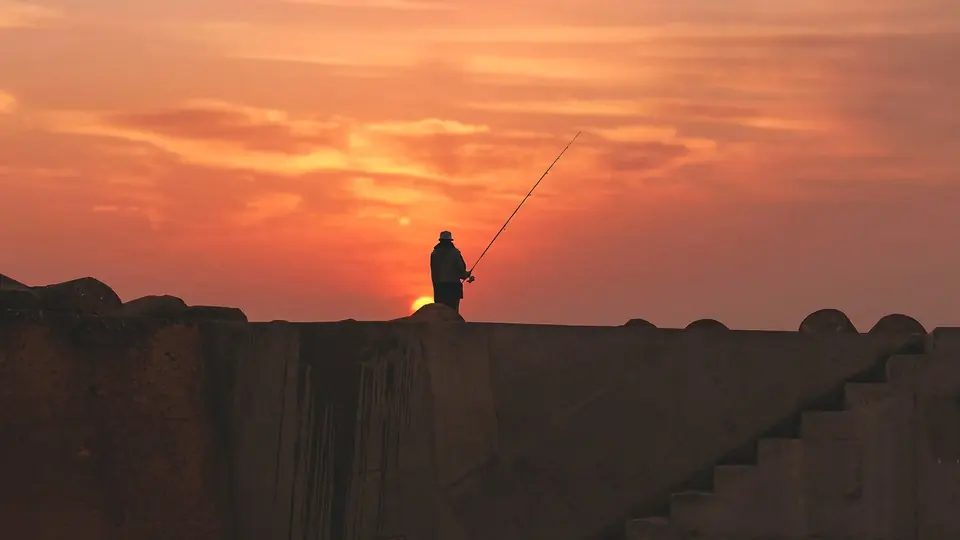 Angler als Silhouette auf einer Hafenmauer vor orangefarbenem Abendhimmel